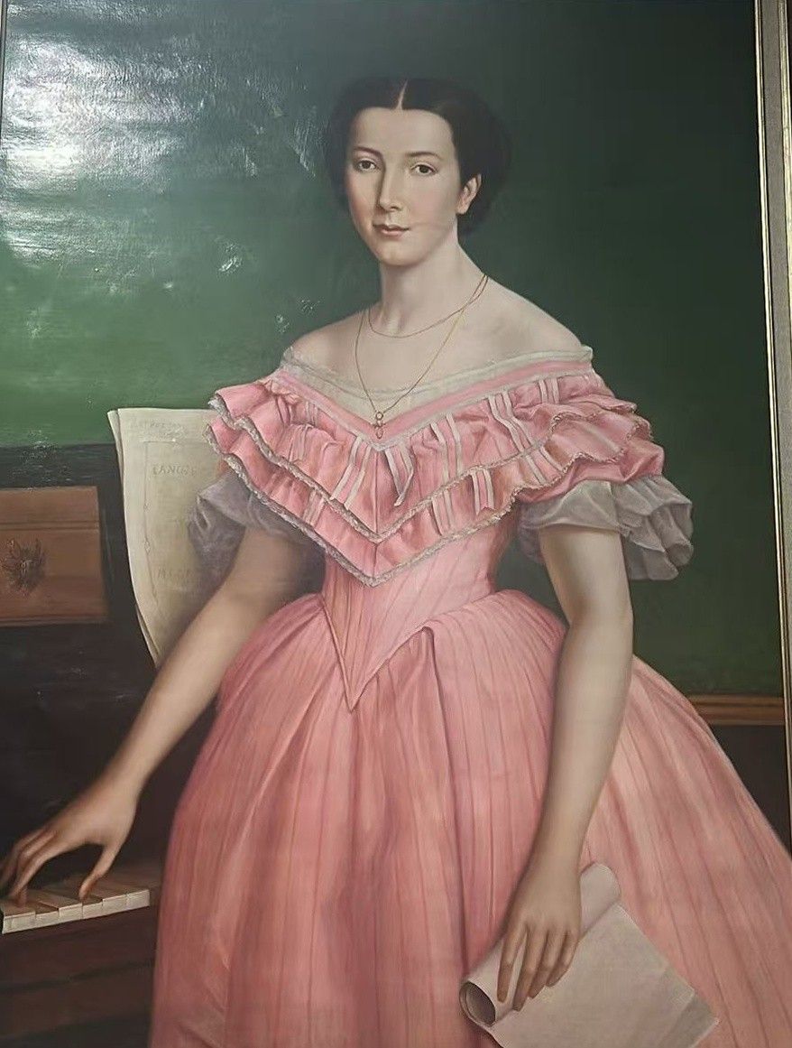 A seated woman in a pink dress poses against a deep green background with books and a writing desk.